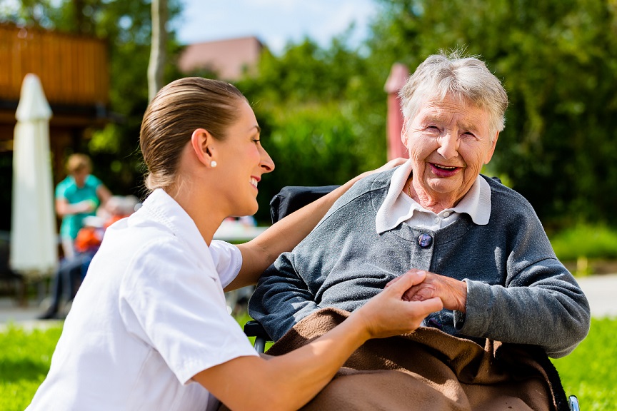 Female caregiver kneels next to elderly woman in a chair while holding her hand